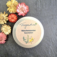 Lade das Bild in den Galerie-Viewer, Geschlossene Metalldose mit dem Produkt „Fragantura – Näschenzauber Nasenbalsam“ auf dunklem Untergrund. Das Etikett zeigt Pflanzenillustrationen und die Produktbezeichnung in blauer Schrift. Links neben der Dose liegen getrocknete Zierblüten in verschiedenen Farben (Gelb, Rosa, Rot, Beige).
