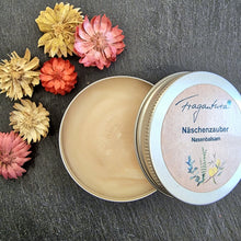 Lade das Bild in den Galerie-Viewer, Runde, silberne Metalldose mit geöffnetem Deckel auf dunklem Untergrund. In der Dose befindet sich ein beigefarbener Nasenbalsam. Auf dem Deckel ist ein Etikett mit der Aufschrift „Fragantura – Näschenzauber Nasenbalsam“ sowie einer Illustration mit zarten Pflanzenmotiven. Links neben der Dose liegen kleine getrocknete Blüten in Gelb, Orange, Rosa und Rot.