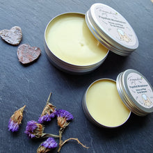 Lade das Bild in den Galerie-Viewer, Das Bild zeigt zwei geöffnete runde Dosen des Fragantura Popoflott Windelbalsams auf dunklem Untergrund. Der gelbe Balsam ist glatt zu sehen. Daneben liegen getrocknete violette Blüten und kleine Herzstücke aus Holz.
