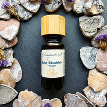 Lade das Bild in den Galerie-Viewer, Kleine Flasche Baby-Bäuchlein Pflegeöl von Fragantura mit naturfarbenem Holzdeckel. Die Flasche ist mittig platziert, umgeben von dekorativen Trockenblumen und grauen Herzsteinen auf dunklem Hintergrund. Das Etikett zeigt dieselbe Baby-in-Hand-Illustration.