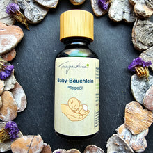 Lade das Bild in den Galerie-Viewer, Braune Glasflasche mit hellbraunem Holzdeckel, etikettiert mit Fragantura Baby-Bäuchlein Pflegeöl. Auf dem Etikett ist eine gezeichnete Hand mit einem entspannt liegenden Baby abgebildet. Die Flasche steht auf schwarzem Untergrund, umrahmt von Trockenblumen und herzförmigen Steinen.