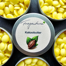 Lade das Bild in den Galerie-Viewer, Runde Metalldose mit Etikettaufschrift „Kakaobutter“ von Fragantura, in der Mitte ein gezeichnetes Kakaoblatt mit Frucht. Um die geschlossene Dose herum sind mehrere geöffnete Dosen mit gelben Kakaobutterplättchen zu sehen.