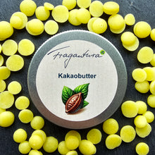Lade das Bild in den Galerie-Viewer, Metalldose mit Etikettaufschrift „Kakaobutter“ von Fragantura liegt inmitten verstreuter gelber Kakaobutterplättchen auf dunklem Untergrund. Auf dem Etikett ist eine Kakaofrucht mit zwei Blättern abgebildet.