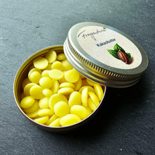 Lade das Bild in den Galerie-Viewer, Geöffnete Metalldose mit gelben Kakaobutterplättchen, daneben der aufgelegte Deckel mit dem Etikett „Kakaobutter“ und einer Zeichnung eines Kakaoblattes samt Frucht. Hintergrund aus dunklem Stein.