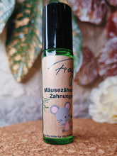 Lade das Bild in den Galerie-Viewer, Ein kleines, grünes Glasfläschchen mit schwarzem Roll-on-Verschluss steht auf einer Korkoberfläche. Das Etikett zeigt die Aufschrift „Fragantura – Mäusezähnchen Zahnungsöl“. Darunter ist eine Illustration einer kleinen grauen Maus mit rosa Ohren zu sehen. Im Hintergrund sind unscharf große, grün-rötliche Blätter sowie eine weiße Blume zu erkennen.