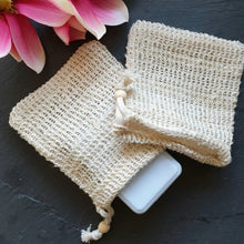 Lade das Bild in den Galerie-Viewer, Ein naturfarbenes, grobmaschiges Seifensäckchen aus Sisal liegt auf dunklem Untergrund. Aus dem offenen Säckchen ragt eine weiße Seife heraus. Links oben im Bild sind rosafarbene Blüten zu sehen.