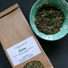 Lade das Bild in den Galerie-Viewer, Papiertüte Fragantura Stilltee neben einer grünen Schale mit losem Kräutertee auf dunklem Hintergrund.