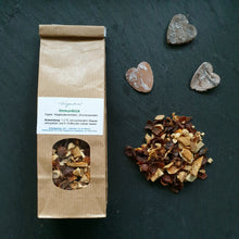 Lade das Bild in den Galerie-Viewer, Braune Papiertüte mit Sichtfenster zeigt grobe Teemischung. Auf dem Etikett steht „Fragantura Immunkick“. Daneben lose Teemischung und drei graue Dekoherzen auf dunklem Untergrund.