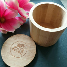 Lade das Bild in den Galerie-Viewer, Eine leere, geöffnete Bambusdose mit Deckel liegt auf dunklem Hintergrund. Der Deckel ist zur Seite gelegt und zeigt eine florale Gravur. Links im Bild sind rosa Magnolienblüten dekorativ platziert.