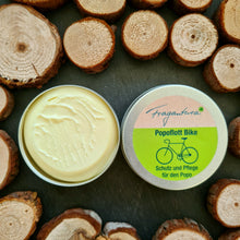 Lade das Bild in den Galerie-Viewer, Das Bild zeigt eine geöffnete Dose Popoflott Bike Creme von Fragantura mit zart gelber, cremiger Oberfläche. Daneben der Deckel, dekoriert mit Baumscheiben auf dunklem Untergrund.