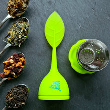 Lade das Bild in den Galerie-Viewer, Zerlegtes grünes Teeei mit Silikonblattgriff und Edelstahleinsatz, daneben drei Teelöffel mit verschiedenen losen Teemischungen – unter anderem mit Kräutern, Lavendelblüten und Fruchtstückchen – auf dunklem Untergrund.