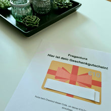 Lade das Bild in den Galerie-Viewer, Abfotografierter Gutschein von Fragantura über 10 Euro, gedruckt auf weißem Papier. In der Mitte ist ein grafisches Geschenk mit roter Schleife und dem Eurozeichen zu sehen. Darunter befindet sich ein verdeckter Gutscheincode. Im Hintergrund steht eine schwarze Dekoschale mit Glaswindlichtern und künstlichen Sukkulenten auf weißem Tisch.
