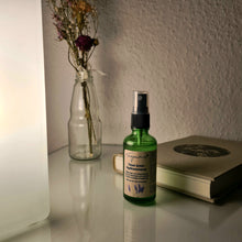 Lade das Bild in den Galerie-Viewer, Ein grünes Glasfläschchen mit Zerstäuberaufsatz steht auf einem Nachttisch, daneben ein geschlossenes Buch und eine Glasvase mit Trockenblumen. Links im Bild ist eine dimmende Lampe zu sehen. Das Etikett trägt die Aufschrift „Träum Schön Kopfkissenspray“ und verweist auf einen Duft aus Zirbelkiefer, Lavendel und Orange. Die Szene wirkt ruhig und entspannend.