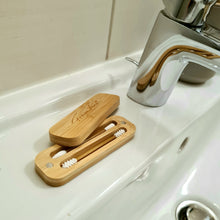 Lade das Bild in den Galerie-Viewer, Ein wiederverwendbares WattestĂ€bchen-Set aus Bambus mit zwei weiĂen SilikonstĂ€bchen liegt geöffnet auf einem weiĂen Waschbecken neben einem Wasserhahn aus Chrom. Auf dem Bambusdeckel ist der Schriftzug âFraganturaâ eingraviert.