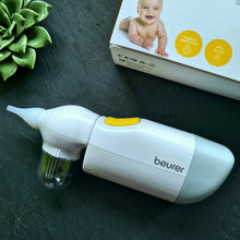 Lade das Bild in den Galerie-Viewer, Elektrischer Nasensauger „Beurer“ mit gelbem Einschaltknopf und durchsichtigem Auffangbehälter, daneben ein Karton mit Babyfoto und gelben Symbolen für hygienische Anwendung und einfache Bedienung. Das Gerät liegt auf dunkler Fläche mit Dekoration aus Sukkulenten.