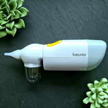 Lade das Bild in den Galerie-Viewer, Weiß-graues elektrisches Gerät der Marke „Beurer“ mit gelbem Knopf, flach liegend auf dunklem Untergrund. Es handelt sich um einen Nasensauger mit durchsichtigem Auffangbehälter vorne. Um das Gerät sind dekorativ grüne künstliche Sukkulenten platziert.