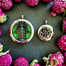 Lade das Bild in den Galerie-Viewer, Zwei runde Duftmedaillons auf dunklem Untergrund mit pinken Trockenblumen: links ein silbernes Medaillon mit Hamsa-Hand und bunten Schmucksteinen auf grünem Filzhintergrund, rechts ein kleineres Medaillon mit ausgestanztem Muster aus Sternen und Herzen.