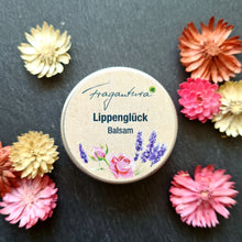 Lade das Bild in den Galerie-Viewer, Die geschlossene Dose mit dem Etikett "Fragantura Lippenglück Balsam" liegt mittig auf dunklem Stein. Um die Dose sind Trockenblumen in Rosa, Rot und Creme verteilt.