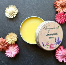 Lade das Bild in den Galerie-Viewer, Das Bild zeigt eine geöffnete Metalldose mit hellem Balsam der Marke Fragantura. Auf dem Deckel steht "Lippenglück Balsam" mit Rosen- und Lavendelmotiv. Rundherum liegen Trockenblumen in Rosa, Rot und Creme.