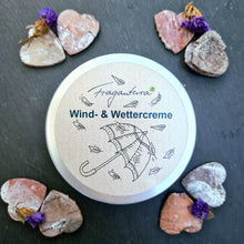 Lade das Bild in den Galerie-Viewer, Eine geschlossene Metalldose der Fragantura Wind- & Wettercreme liegt mittig, umrahmt von sechs herzförmigen Holzstücken mit Trockenblumen. Der Deckel zeigt einen Regenschirm mit fallenden Blättern und die Aufschrift „Wind- & Wettercreme“. Der Hintergrund ist aus dunklem Schieferstein.