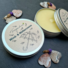 Lade das Bild in den Galerie-Viewer, Eine geöffnete Metalldose zeigt die gelbe Wind- & Wettercreme von Fragantura. Daneben liegt der bedruckte Deckel mit Regenschirm-Illustration und Schriftzug „Wind- & Wettercreme“. Um die Dose sind getrocknete Blüten und Rindenherzen dekorativ arrangiert.