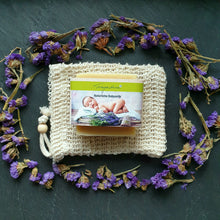Lade das Bild in den Galerie-Viewer, Eine rechteckige, naturfarbene Seife mit Etikett "Fragantura - Natürliche Babyseife" liegt auf einem beigen Sisalsäckchen. Um das Säckchen herum ist ein Kreis aus getrockneten, violetten Blütenblättern arrangiert.