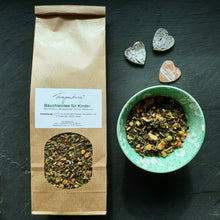 Lade das Bild in den Galerie-Viewer, Aufrecht liegende Tüte „Bäuchleintee für Kinder“ neben einer Schale mit Teemischung und zwei kleinen Dekoherzen aus Holz. Der Tee ist in einer transparenten Sichtöffnung der Tüte sichtbar. Die Schale enthält dieselbe Kräutermischung und steht rechts daneben.