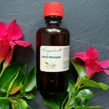 Lade das Bild in den Galerie-Viewer, Braune Glasflasche mit rotem Deckel, etikettiert mit „Fragantura Damm-Massageöl“. Auf dem Etikett ist eine Lilie abgebildet. Die Flasche liegt auf grauem Untergrund, umrahmt von frischen roten Blumen mit grünen Blättern.