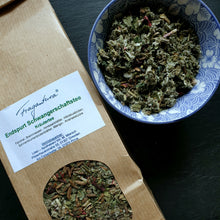 Lade das Bild in den Galerie-Viewer, Nahaufnahme des Etiketts auf der Teeverpackung mit deutlich lesbaren Zutaten wie Fenchel, Brennnessel und Himbeerblättern, daneben eine geöffnete Teeschale mit getrockneten Kräutern.