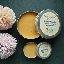 Lade das Bild in den Galerie-Viewer, Zwei geöffnete Metalldosen mit goldgelber Goldröschensalbe von Fragantura, jeweils mit Deckel daneben. Die größere Dose trägt die Aufschrift: „Fragantura – Goldröschensalbe – Linderung und Pflege für wunde Hautpartien“. Dekoriert mit zwei pastellfarbenen Blüten auf dunklem Hintergrund.