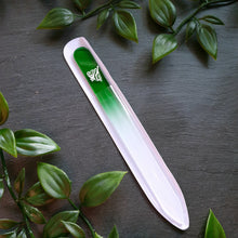 Lade das Bild in den Galerie-Viewer, Glasnagelfeile mit Farbverlauf von Dunkelgrün zu Weiß, Liliengravur und transparenter Schutzhülle, umgeben von grünen Kunstblättern auf schwarzem Untergrund.