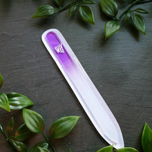 Lade das Bild in den Galerie-Viewer, Glasnagelfeile mit Farbverlauf von Violett zu Weiß, eingraviertem Liliensymbol und transparenter Hülle, präsentiert auf dunklem Untergrund mit grüner Blätterdekoration.