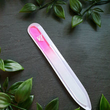 Lade das Bild in den Galerie-Viewer, Glasnagelfeile mit Farbverlauf von Rosa zu Weiß, eingraviertem Lilienmotiv und transparenter Schutzhülle, dekoriert mit grünen Kunstpflanzen auf dunklem Hintergrund.