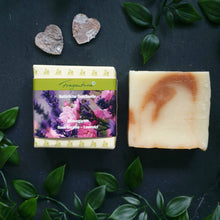 Lade das Bild in den Galerie-Viewer, Das Bild zeigt ein rechteckiges Seifenstück der Marke Fragantura neben der dazugehörigen Verpackung. Die Banderole trägt die Aufschrift „Fragantura – Natürliche Duschseife – Blütengarten – Rose Geranie – Lavendel“ und zeigt florale Motive in Rosa und Violett. Die Szene ist mit grünen Blättern und zwei kleinen Herzdekorationen gestaltet.