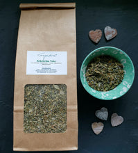 Lade das Bild in den Galerie-Viewer, Vollansicht der Teetüte mit Etikett auf dunklem Hintergrund, daneben die grüne Schale mit Tee und drei kleine Steinherzen als Dekoration.
