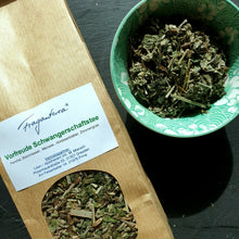 Lade das Bild in den Galerie-Viewer, Zu sehen ist ein Teil der Teeverpackung aus braunem Kraftpapier mit dem Etikett „Vorfreude Schwangerschaftstee“ sowie eine grüne Porzellanschale mit stilisiertem Blütenmuster, gefüllt mit dem losen Kräutertee. Die Mischung enthält gut erkennbare Blätter, Stängel und Pflanzenteile. Das Bild ist bei Tageslicht aufgenommen und vermittelt eine natürliche, ruhige Stimmung.