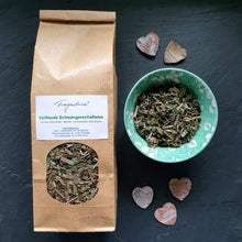 Lade das Bild in den Galerie-Viewer, Das Bild zeigt eine braune Kraftpapier-Verpackung des Kräutertees „Vorfreude Schwangerschaftstee“ von Fragantura mit Sichtfenster. Rechts daneben steht eine grüne Schale mit floralem Muster, gefüllt mit grob geschnittenem Kräutertee. Auf dem Etikett sind Zutaten wie Fenchel, Brennnessel und Zitronengras aufgeführt. Drei kleine Deko-Herzen aus Holz liegen daneben auf dunklem Schieferuntergrund.