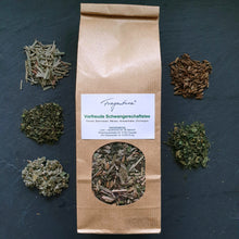 Lade das Bild in den Galerie-Viewer, Die braune Teeverpackung „Vorfreude Schwangerschaftstee“ von Fragantura steht mittig auf dunklem Schiefer. Rundherum sind die getrockneten Einzelzutaten in kleinen Häufchen angeordnet: Zitronengras, Melisse, Himbeerblätter, Brennnessel und Fenchelsamen. Das Sichtfenster der Verpackung gibt den Blick auf die gemischte Kräuterkomposition frei.