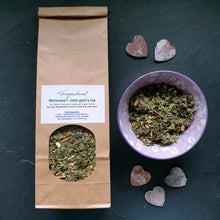 Lade das Bild in den Galerie-Viewer, Eine braune Papiertüte mit Sichtfenster zeigt den losen Kräutertee „Wehentee – Jetzt geht’s los“ der Marke Fragantura. Auf dem weißen Etikett sind Zutaten wie Zimt, Nelke, Eisenkraut, Liebstöckel, Ingwer und Beifuß aufgeführt, mit dem Hinweis: „Nur nach Rücksprache mit Ihrer Hebamme anwenden!“. Rechts daneben steht eine violett gemusterte Keramikschale mit der trockenen Teemischung. Der Hintergrund ist eine dunkle Schieferplatte, dekoriert mit kleinen Holzherzen.