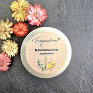 Geschlossene Metalldose mit dem Produkt „Fragantura – Näschenzauber Nasenbalsam“ auf dunklem Untergrund. Das Etikett zeigt Pflanzenillustrationen und die Produktbezeichnung in blauer Schrift. Links neben der Dose liegen getrocknete Zierblüten in verschiedenen Farben (Gelb, Rosa, Rot, Beige).