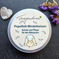 Das Bild zeigt eine geschlossene runde Dose Fragantura Popoflott Windelbalsam auf dunklem Stein. Auf dem Etikett sind ein Baby im Windelanzug und weiße Federn abgebildet. Violette Blüten und ein silbernes Herz liegen daneben.