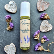 Ein einzelner Roll-On der Duftserie „Träum Schön“ von Fragantura liegt auf dunklem Schieferuntergrund. Das kleine Glasfläschchen mit weißem Deckel trägt ein naturfarbenes Etikett mit Lavendelmotiv und dem Aufdruck „Träum Schön – Roll-On“. Rund um das Produkt liegen getrocknete violette Blütenköpfe sowie mehrere kleine herzförmige Steine als liebevolle Dekoration.