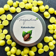 Metalldose mit Etikettaufschrift „Kakaobutter“ von Fragantura liegt inmitten verstreuter gelber Kakaobutterplättchen auf dunklem Untergrund. Auf dem Etikett ist eine Kakaofrucht mit zwei Blättern abgebildet.