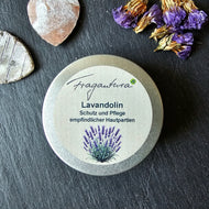 Runde silberne Metalldose mit Etikett „Fragantura Lavandolin – Schutz und Pflege empfindlicher Hautpartien“, darunter eine Illustration von Lavendel. Daneben liegen lila Trockenblüten und zwei rustikale Herzdekorationen auf dunklem Untergrund.