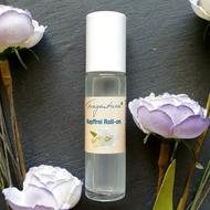Klare Glasflasche mit weißem Deckel, beschriftet mit dem Etikett „Kopffrei Roll-on“ der Marke Fragantura. Auf dem Etikett befindet sich eine weiße Lilienblüte. Das Produkt steht auf dunklem Untergrund, umrahmt von dekorativen Stoffblüten in Weiß und Lavendel.
