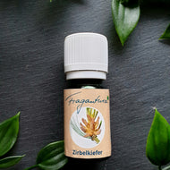 Das Bild zeigt eine kleine grüne Glasflasche mit weißem Schraubverschluss, zentral auf dunklem Schieferhintergrund platziert. Die Flasche trägt ein Etikett mit der Aufschrift „Fragantura Zirbelkiefer“. Auf dem Etikett ist eine Illustration eines frischen Zirbelkiefer-Zapfens mit Nadeln abgebildet. Rund um die Flasche liegen dekorativ grüne Blätter verteilt. Es handelt sich um ein ätherisches Öl mit Zirbelkieferduft, das typischerweise zur Raumbeduftung verwendet wird.