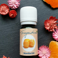 Braunes Glasfläschchen mit weißem Schraubverschluss, beschriftet mit „Fragantura – Mandarine“. Auf dem Etikett ist ein Bild von zwei geschälten Mandarinen zu sehen. Die Flasche steht auf dunklem Schieferuntergrund und ist umgeben von orangenen Mandarinenstücken und dekorativen rosa bis roten Trockenblumen.