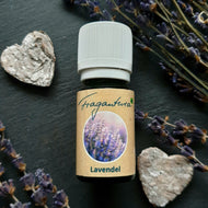 Kleine grüne Glasflasche mit weißem Schraubverschluss, beschriftet mit dem Etikett „Fragantura Lavendel“. Auf dem Etikett befindet sich ein rundes Bild von blühendem Lavendel. Die Flasche liegt auf dunklem Schiefer, dekoriert mit getrockneten Lavendelblüten und zwei rustikalen Holzherzen.