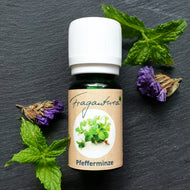 Kleine grüne Flasche Fragantura Pfefferminze-Öl mit weißem Deckel, dekoriert mit frischer Minze und violetten Blüten auf dunklem Stein.