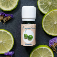Kleine grüne Glasflasche mit weißem Schraubverschluss, Etikett beschriftet mit „Fragantura Limette“ und einem Bild von zwei grünen Limetten. Die Flasche liegt auf dunklem Untergrund, umgeben von frischen Limettenscheiben und violetten Trockenblüten.